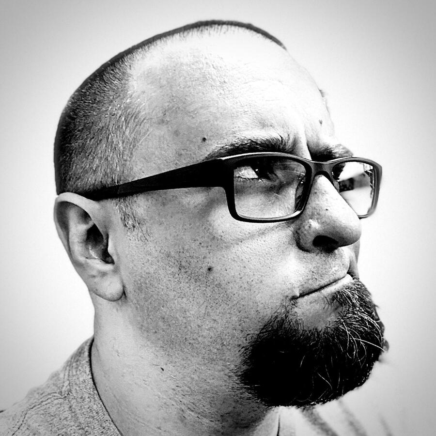 Black & white portrait of a man with very short hair and a thick but short goatee. He is wearing black glasses and a gray t-shirt and looking to his left with an expression like he's not sure about something.